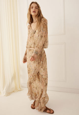 A woman modeling a long-sleeved maxi dress with a beige and brown paisley pattern, standing in a bright, minimalist room with white walls and a cream curtain.